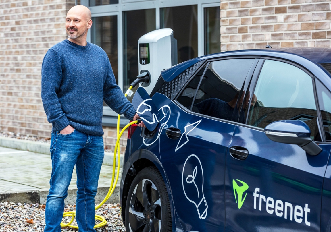 Ein Mann lädt ein blaues Elektroauto an einer Ladestation auf, das Auto trägt das freenet-Logo und Symbole für Nachhaltigkeit.