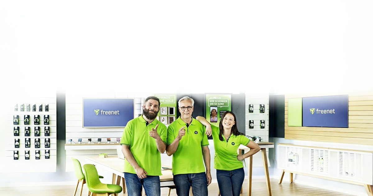 Drei Mitarbeiter in grünem Freenet-Poloshirt lächeln in einem modernen Laden mit Freenet-Logo und Zubehör.